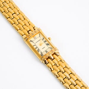May include: A gold-tone rectangular wristwatch with a link bracelet. The cream-coloured dial features black Roman numeral hour markers, gold hands, and the text LANCEL PARIS. Small clear stones are set into the bezel above and below the dial.