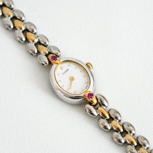 May include: A Tisse wristwatch featuring an oval silver-tone case with a white dial and gold-tone hands. The watch band consists of alternating silver and gold-tone oval links. Two small pink stones are set at the top and bottom of the watch case.