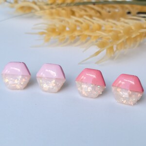 May include: Four pairs of pink and white hexagonal stud earrings with iridescent glitter. The earrings are arranged in a row on a white surface.