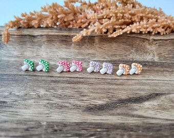 May include: A collection of colorful mushroom-shaped stud earrings. Each pair features a different color cap, including green, pink, purple, and orange, with white polka dots. The earrings have a white stem and are displayed on a wooden surface.