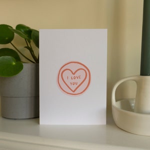 Può includere: Un biglietto bianco con un cuore rosa e il testo "I love you" su di esso. Il biglietto è appoggiato su una superficie bianca accanto a un portacandele bianco e una candela verde.