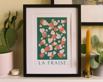 Può includere: Stampa artistica incorniciata con un'illustrazione botanica di fragole e foglie su sfondo verde scuro. La stampa è in una cornice nera e presenta il testo "LA FRAISE" in basso. Sono visibili anche un cactus in un vaso rosa e delle candele.