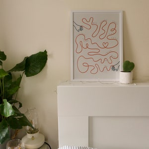 Pu&ograve; includere: Una stampa incorniciata bianca con un disegno a linea rossa di mani che si raggiungono a vicenda. Il testo "We all have the strength to grow" &egrave; scritto in nero all'interno del disegno. La stampa &egrave; appesa a una testata bianca sopra un cuscino a righe bianche e blu.