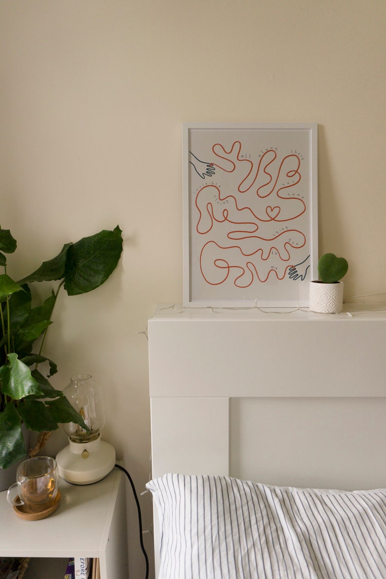 Pu&ograve; includere: Una stampa incorniciata bianca con un disegno a linea rossa di mani che si raggiungono a vicenda. Il testo "We all have the strength to grow" &egrave; scritto in nero all'interno del disegno. La stampa &egrave; appesa a una testata bianca sopra un cuscino a righe bianche e blu.