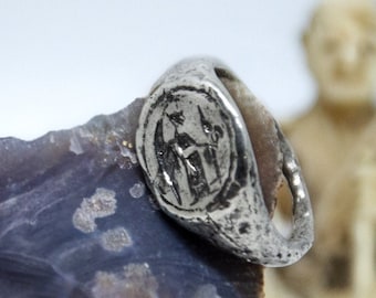 May include: A silver ring with a raised design of two figures standing side by side. The ring is set on a purple and gray stone.