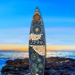 May include: A surfboard shaped mosaic art piece with a blue, green, and brown color scheme. The mosaic is made of small tiles and features a variety of patterns, including circles, swirls, and waves. The surfboard is set against a backdrop of a beach scene with blue water and a sandy shore.
