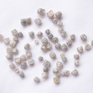May include: A collection of small, grey, rough-cut diamonds, also known as industrial diamonds, scattered on a white surface.