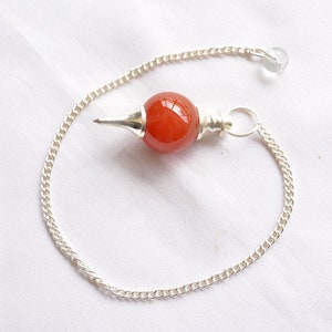 Könnte beinhalten: Eine silberne Kette mit einem roten Karneol-Kristallpendel. Das Pendel ist rund und hat eine spitze Spitze.