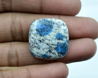 May include: A smooth, oval-shaped cabochon gemstone with a white base and blue flecks. The stone has a speckled appearance.
