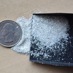 Pode incluir: Um pequeno monte de vidro branco, brilhante e triturado com uma p&aacute; preta e uma moeda de um quarto de d&oacute;lar americano para escala.