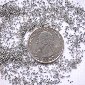 Pode incluir: Um monte de pequenos diamantes cinzentos espalhados sobre uma superf&iacute;cie branca. Uma moeda de um quarto de d&oacute;lar americano &eacute; colocada no centro do monte para servir de escala.