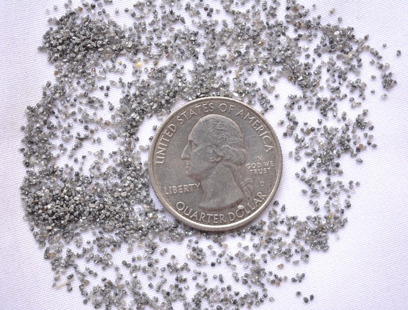 Pode incluir: Um monte de pequenos diamantes cinzentos espalhados sobre uma superf&iacute;cie branca. Uma moeda de um quarto de d&oacute;lar americano &eacute; colocada no centro do monte para servir de escala.
