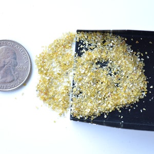 Pode incluir: Um pequeno monte de material amarelo e branco semelhante a glitter ao lado de uma moeda de um quarto de d&oacute;lar americano. O glitter est&aacute; em uma bandeja preta pequena.