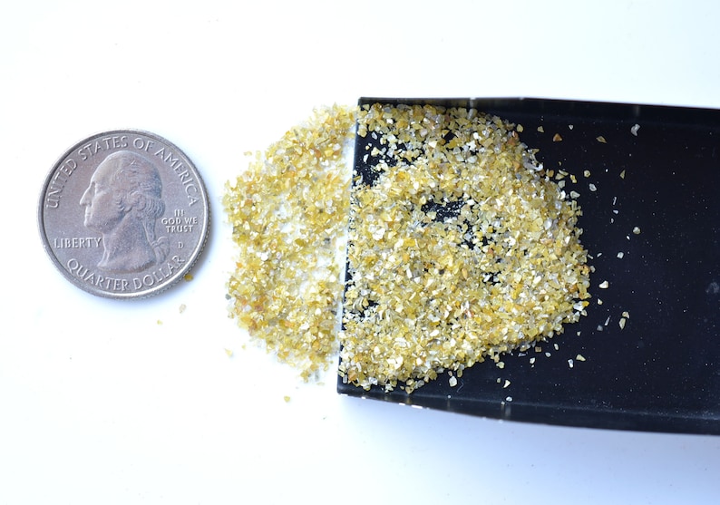 Pode incluir: Um pequeno monte de material amarelo e branco semelhante a glitter ao lado de uma moeda de um quarto de d&oacute;lar americano. O glitter est&aacute; em uma bandeja preta pequena.