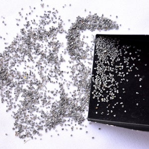 Pode incluir: Uma pequena bandeja preta cheia de diamantes cinzentos soltos. Os diamantes est&atilde;o espalhados sobre uma superf&iacute;cie branca.