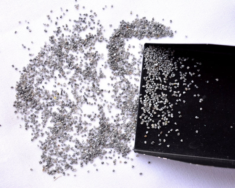 Pode incluir: Uma pequena bandeja preta cheia de diamantes cinzentos soltos. Os diamantes est&atilde;o espalhados sobre uma superf&iacute;cie branca.