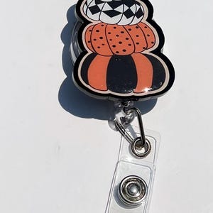 May include: A stacked pumpkin badge reel with a black border. The pumpkins are white, orange, and black with various patterns. The reel has a clear plastic strap and a metal clip.