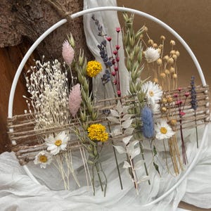 Könnte beinhalten: Ein weißer runder Reifen, verziert mit getrockneten Blumen und Gräsern, darunter gelbe Trommelstöckchen, rosa Hasenschwanzgras, Lavendel und weiße Gänseblümchen. Das Arrangement ist mit horizontalen braunen Garnfäden befestigt.