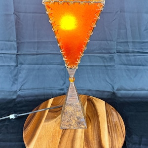 May include: A unique table lamp with a triangular orange shade, stitched with a light-colored thread. The lamp has a wooden base and sits on a round wooden surface. The lamp's design is complemented by a warm, inviting glow.