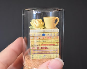 以下が含まれることがあります： コルクの蓋が付いたガラス瓶の中に、ミニチュアの積み重ねられた本が入っています。その上に、黄色のティーカップと黄色の花が置かれています。本には、「動物農場」や「コレラの流行の愛」などのタイトルが書かれています。