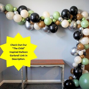 May include: A balloon garland featuring black, silver, white, green, and brown balloons. The garland is hanging on a wall and is decorated with a star-shaped sign that says "Check Out Our "The Child" Inspired Balloon Garland! Link in Description."