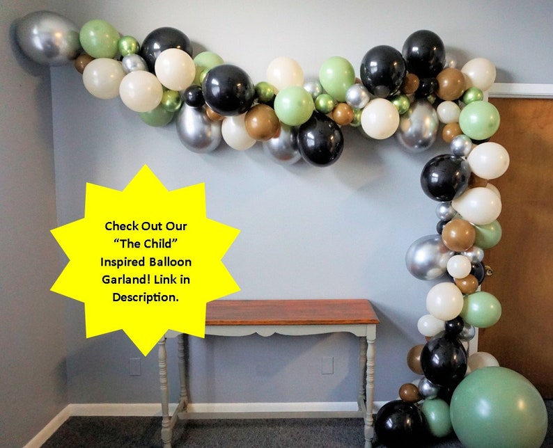 May include: A balloon garland featuring black, silver, white, green, and brown balloons. The garland is hanging on a wall and is decorated with a star-shaped sign that says "Check Out Our "The Child" Inspired Balloon Garland! Link in Description."