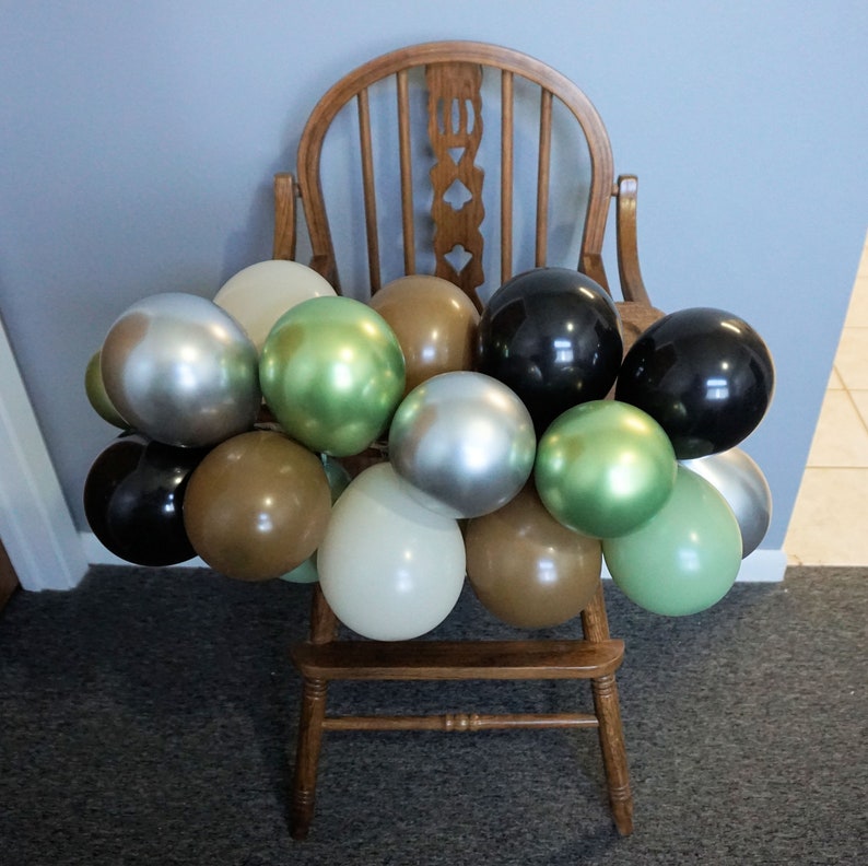 May include: A wooden chair decorated with a balloon garland in shades of brown, green, silver, and black. The balloons are clustered around the chair.