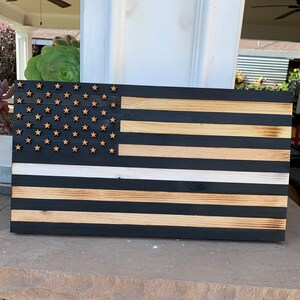 Puede incluir: Una bandera americana de madera con una fina franja blanca en el medio. La bandera está pintada de negro y tiene un acabado de madera natural. Las estrellas son de madera y están pintadas de un color marrón claro.