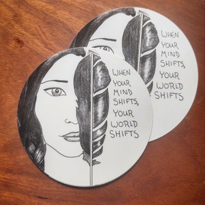 May include: Two black and white circular stickers with a woman's face and a feather. The text on the stickers reads "When your mind shifts, your world shifts."