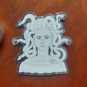May include: A silver sticker featuring a black and white line drawing of Medusa, a mythical Greek figure with snakes for hair. Her eyes are purple and she has a snake wrapped around her neck.