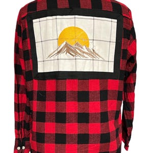 May include: Red and black plaid flannel shirt with a white patch on the back featuring an embroidered mountain scene with a yellow sun.