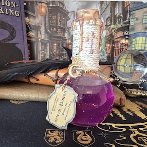 Könnte beinhalten: Eine runde Glasflasche mit violetter Flüssigkeit und Glitzer, deren Hals mit weißem Seil umwickelt und mit goldenen Wachstropfen verziert ist. Ein goldener Mondsichel-Anhänger hängt an einer Kette, dazu ein beiges Etikett mit der Aufschrift Sleep Potion.