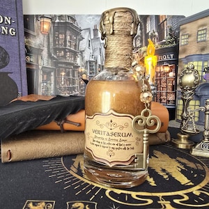 Könnte beinhalten: Eine klare Glasflasche gefüllt mit einer schimmernden goldenen Flüssigkeit und einem Etikett mit der Aufschrift VERITASERUM. Der Flaschenhals ist mit Schnur umwickelt und mit einem goldenen Wachssiegel versehen. Ein goldener Skelettschlüssel hängt am Hals.