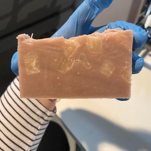 May include: A rectangular bar of handmade soap with a light pink hue. The soap is embedded with small, light-colored, square-shaped pieces. The soap is held by a person wearing a blue glove. The background is a white surface.