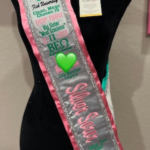 May include: A silver, pink, and green sash with the text "Silver Soror" and "Fisk University" embroidered on it. The sash also includes the Greek letters "Π" and "ΒΕΩ" and the text "Clean, Mean Chosen 15 DOP 1980 Big Sister "Most Gracious" 12-Π-1978 Graduatin Member Lite Member Legacy".