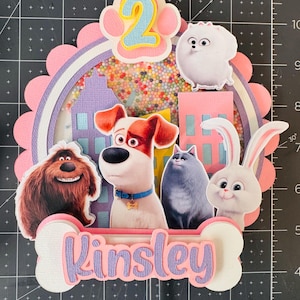 May include: A layered cake topper featuring characters from "The Secret Life of Pets" movie. The design includes Max, Duke, Gidget, Snowball, and a cat. The topper has a pink and purple color scheme, with the name "Kinsley" on a bone-shaped element.