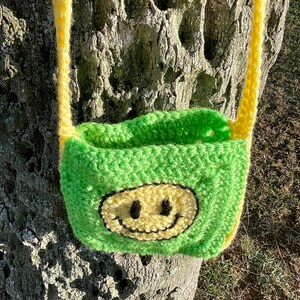 May include: A green crocheted bag with a yellow smiley face design. The bag has yellow straps and is hanging from a tree trunk.
