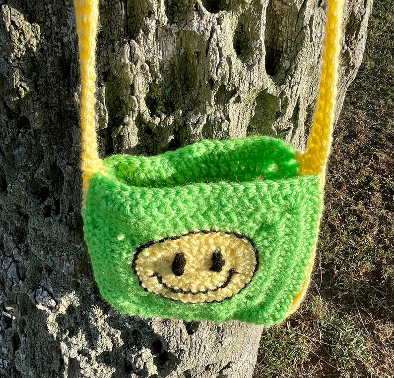 May include: A green crocheted bag with a yellow smiley face design. The bag has yellow straps and is hanging from a tree trunk.