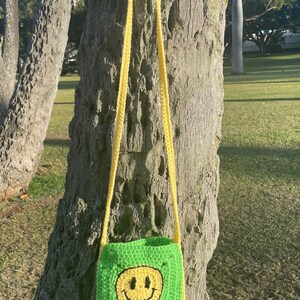 May include: A green crocheted crossbody bag with a yellow smiley face design. The bag is hanging on a tree trunk.