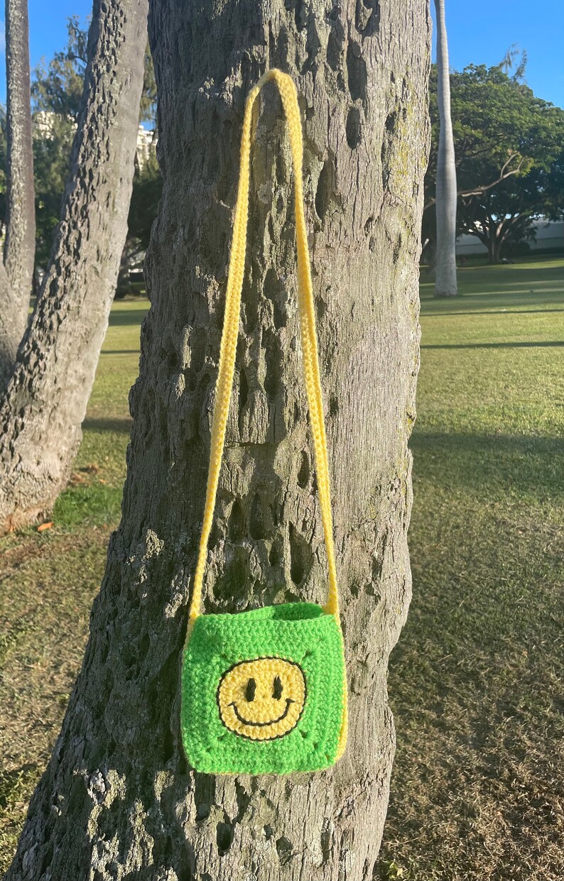 May include: A green crocheted crossbody bag with a yellow smiley face design. The bag is hanging on a tree trunk.