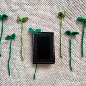 May include: A black book with a gold trim is surrounded by green crocheted plant bookmarks. The bookmarks have a three-leaf clover design.