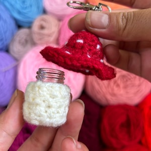Puede incluir: Un hongo rojo de ganchillo con lunares blancos, unido a un llavero plateado, y una cubierta de ganchillo blanca para una pequeña botella de vidrio.