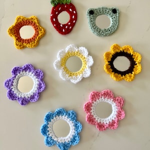 May include: A collection of handmade crochet mirrors in various shapes and colors. Designs include a strawberry, a frog, and several flower shapes in shades of yellow, purple, white, pink, and blue. Each mirror has a reflective surface.