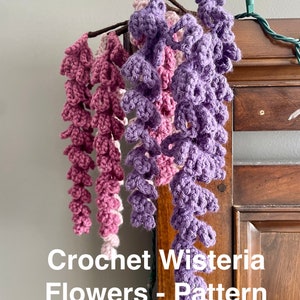 May include: Crocheted wisteria flowers in shades of purple and pink hanging from a wooden branch. The text "Crochet Wisteria Flowers - Pattern Only!!" is written below the flowers.
