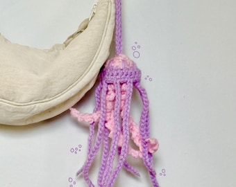 May include: A handmade crochet jellyfish bag charm in shades of pink and purple. The jellyfish has a rounded head and long, flowing tentacles. The charm is attached to a purple cord and is hanging next to a beige bag. Text on the image reads "Crochet Jellyfish Bag Charm."