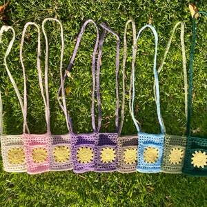 Op de afbeelding: Acht gehaakte mini-tasjes met verstelbare bandjes. Elk tasje heeft een andere kleur en een geel bloemenontwerp.