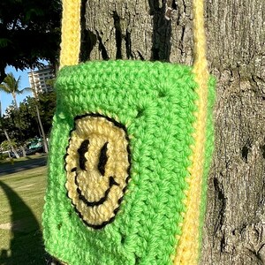 May include: A green and yellow crocheted crossbody bag with a smiley face design. The bag is hanging on a tree trunk.