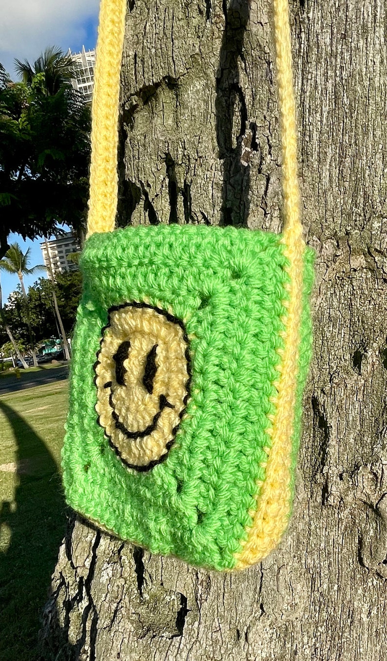 May include: A green and yellow crocheted crossbody bag with a smiley face design. The bag is hanging on a tree trunk.