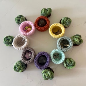 May include: A collection of handmade crochet bowls in various colors, each containing small items like hair ties, coins, and jewelry. The bowls are arranged in a circular pattern, with green and brown decorative accents.