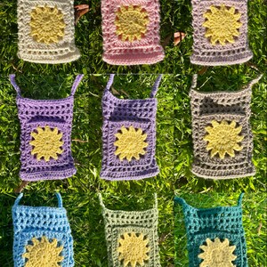 Op de afbeelding: Negen gehaakte tassen met een zonnebloemdessin. De tassen zijn in verschillende kleuren verkrijgbaar, waaronder cr&egrave;me, roze, paars, lichtblauw, groen en turquoise. Elke tas heeft een band en een klein vakje met een gehaakte zonnebloem.